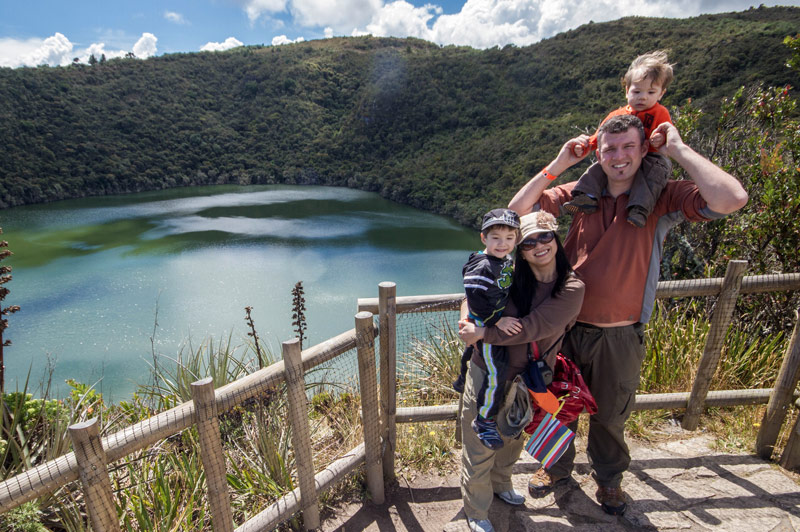 A young family smiles while standing on an over look over a crater lake surrounded by jungle as they search for the lost city of gold in Guatavita Colombia with kids