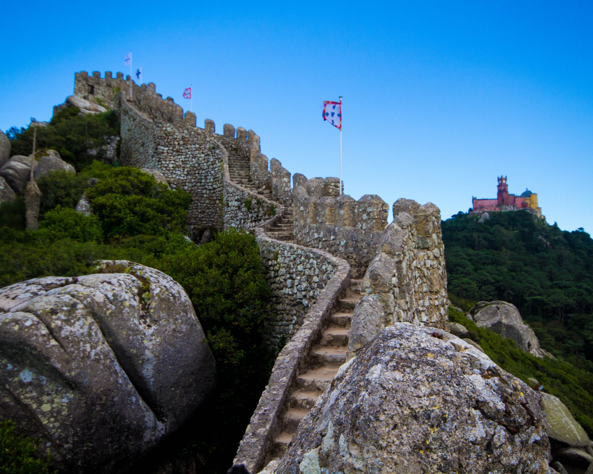 Looking along the wall of a stone castle with a colourful palace in the background - Sintra, Portugal