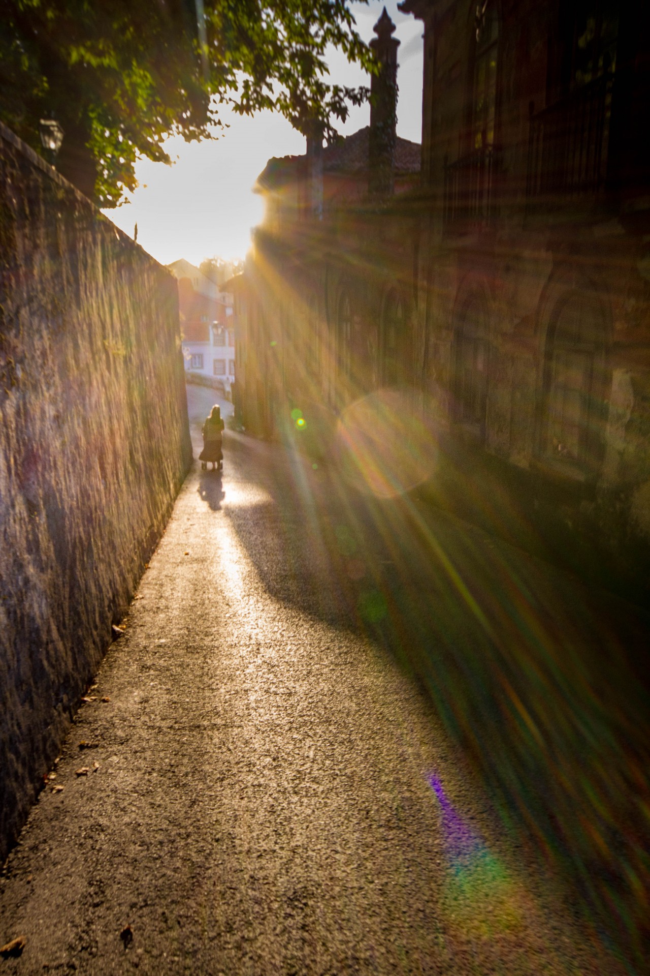 A woman pushes a baby stroller down a narrow cobblestone street while the suns rays explode around her - Sintra, Portugal