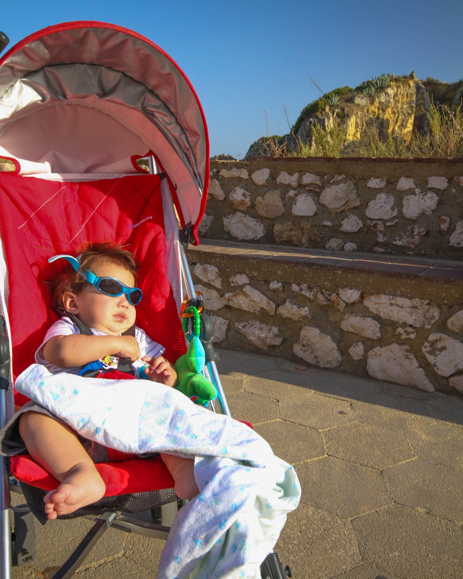 Baby with sunglasses on sits in a stroller at the beach - helping kids find nap time on the road