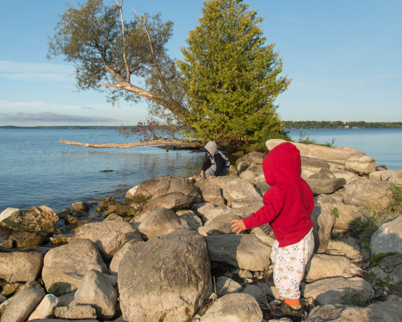 A toddler in a red hoodie and pyjamas climbs over rocks near a lake while camping in Mara Provincial Park