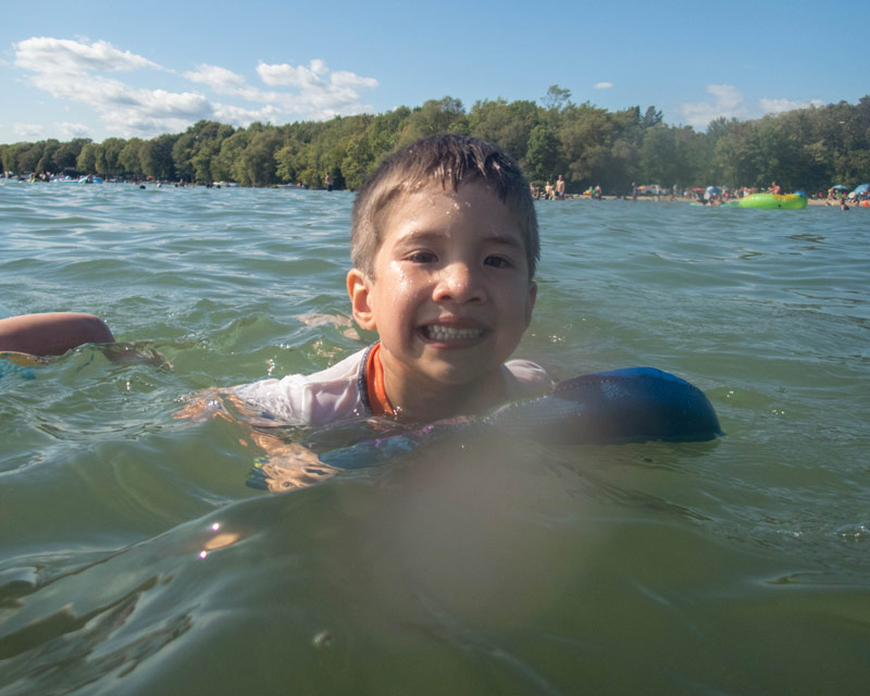 A young boy swims with the help of a board in a lake while camping in Mara Provincial Park with kids