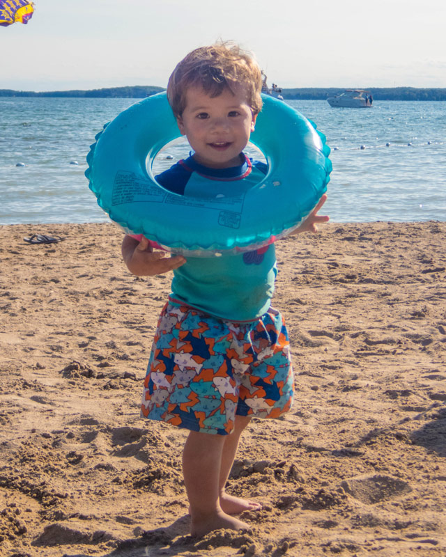 A toddler stands on a beach while holding an inflatable tube like a necklace while camping in Mara Provincial Park with kids