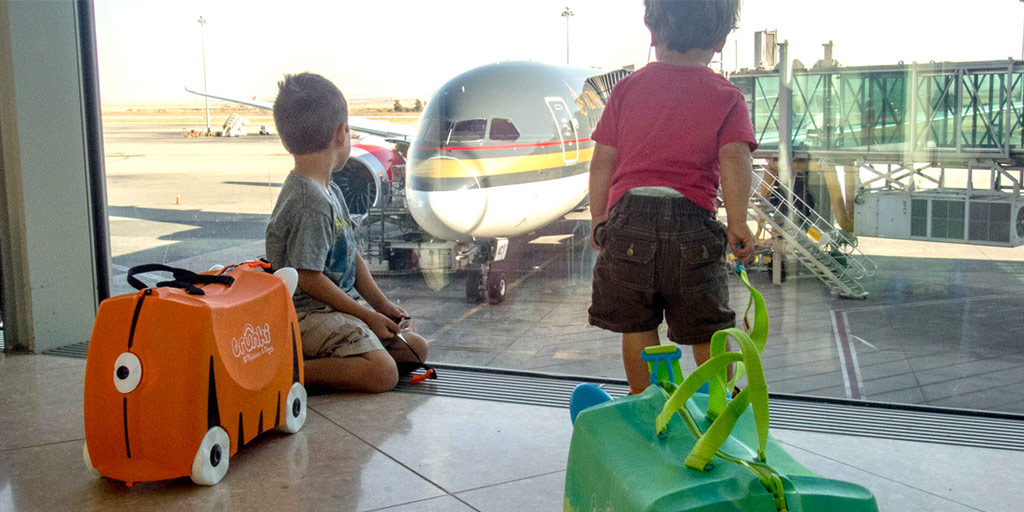 two young boys with cute character luggages look at an airplane through the window of an airport