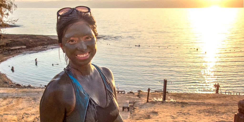 A smiling asian woman covered in mud stands in front of the Dead Sea in Jordan at sunset