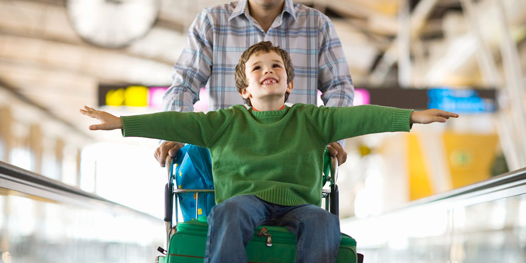 A smiling boy sitting on a luggage cart is pushed through an airport by his father