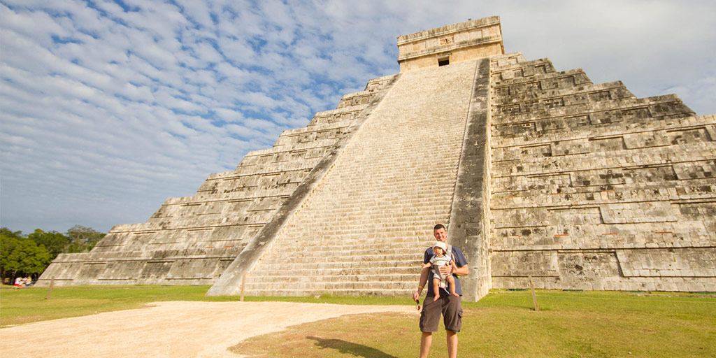 a man holding a baby stands beside a large mayan pyramid in Mexico