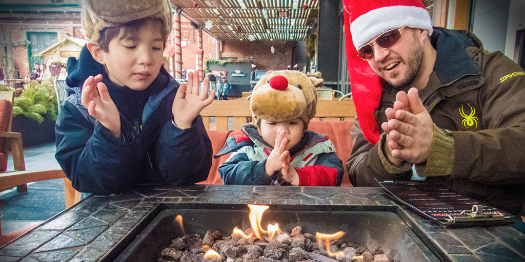 A man wearing a Santa hat and two young boys wearing rudolph hats warm themselves by a fire at a Christmas Market