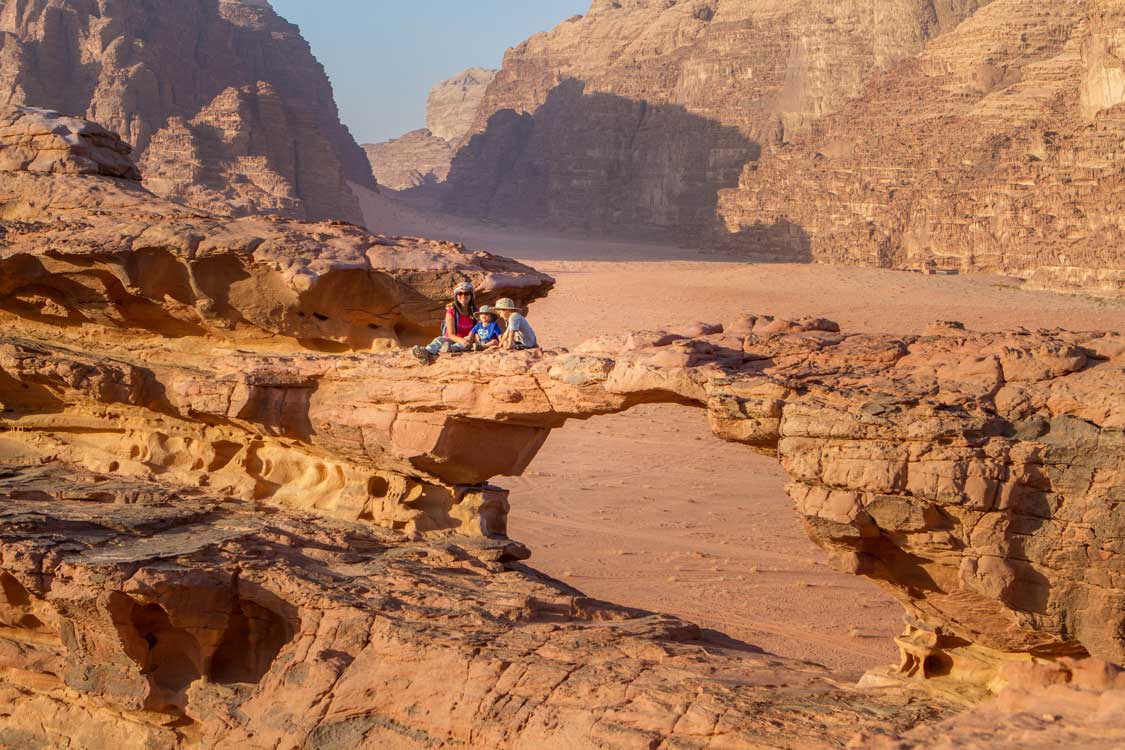 A woman and two children sit on a rock arch in a Jordanian desert