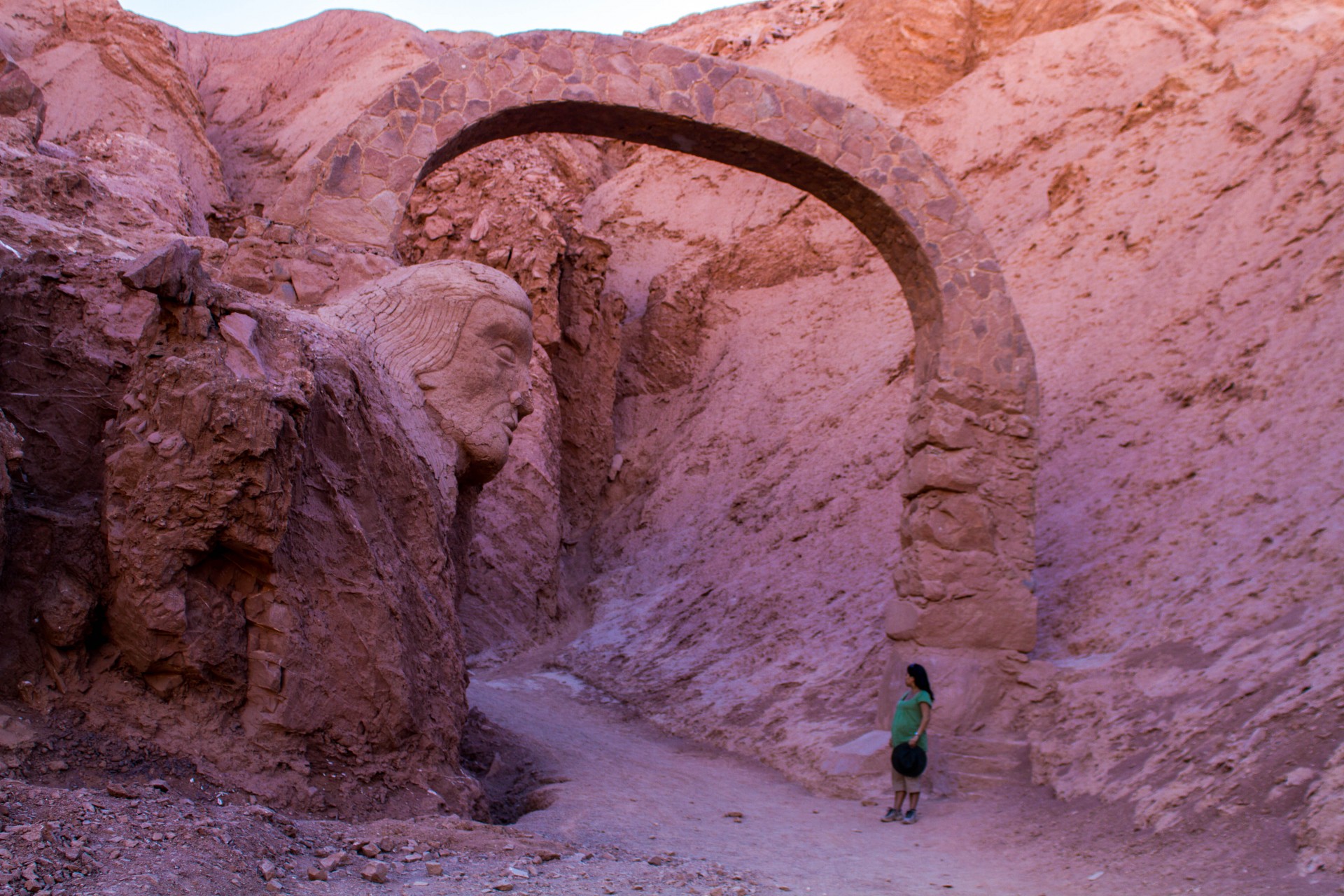 A pregnant woman stands near a tall stone arch with a carving of an indigenous person