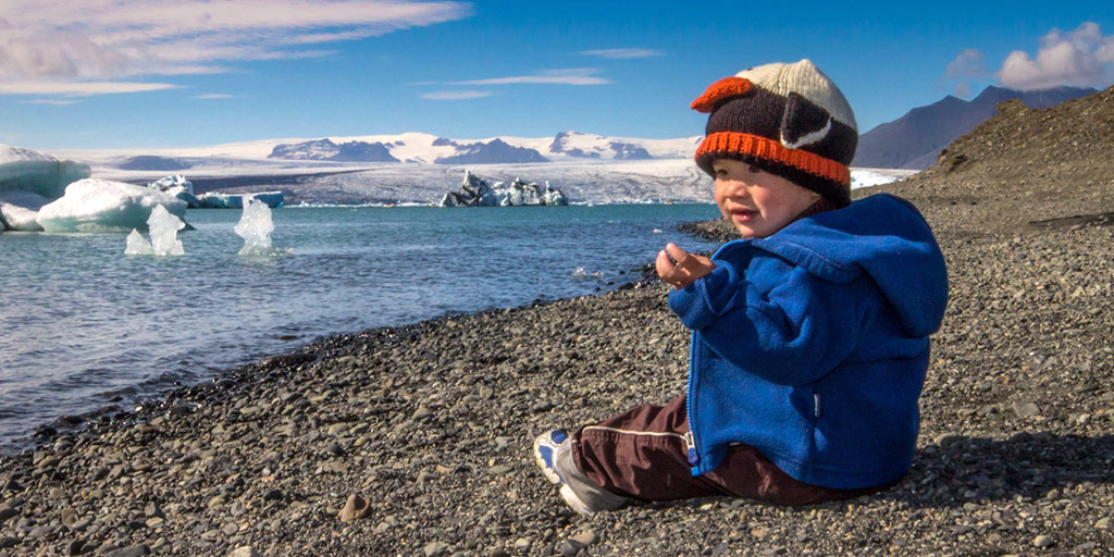 Young boy in a penguin hat sitting on a rocky beach looking out at glaciers and icebergs