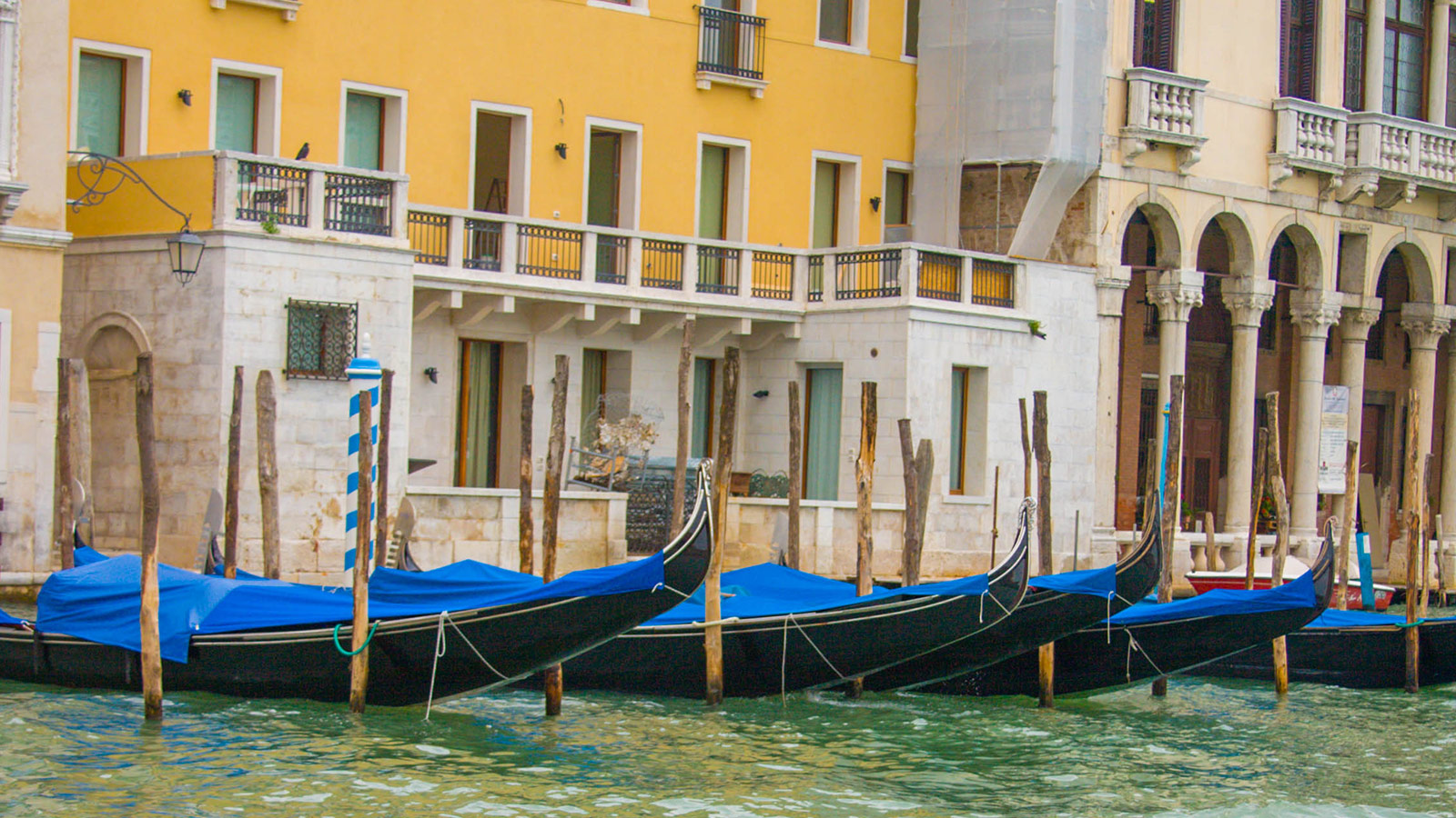 Venetial gondolas covered in blue sit docked against a yellow building