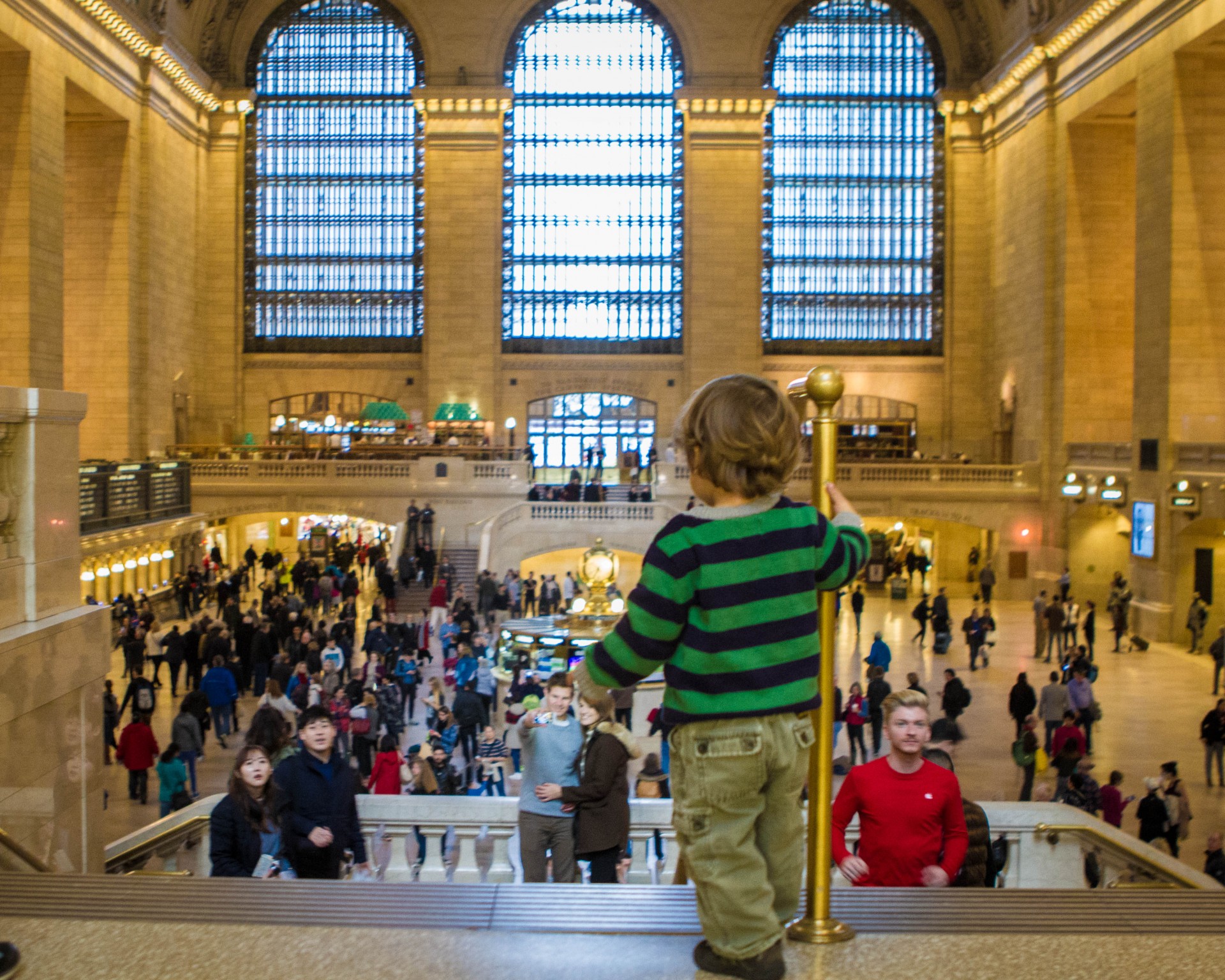 A toddler stands at the top of the stairs looking out over the inside of Grand Central Station in New York City