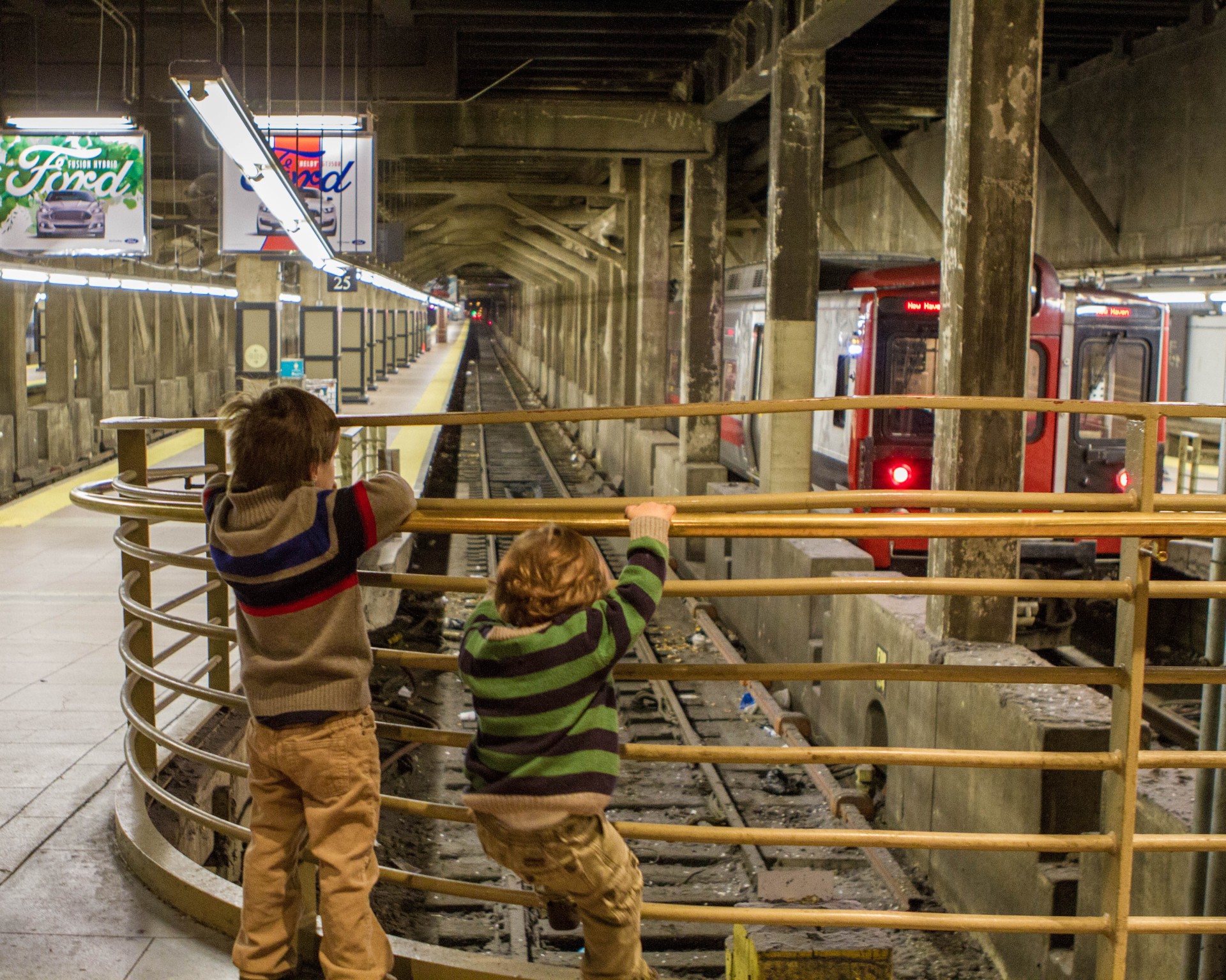 two young boys hang on to a gate while looking at a subway station tracks