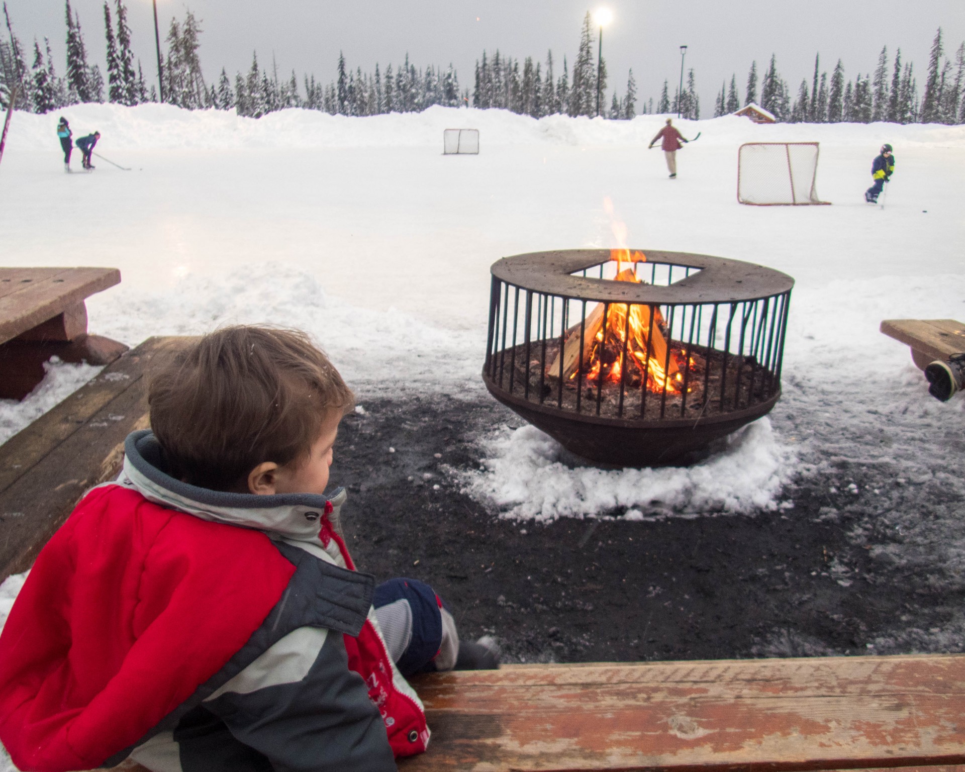 Young boy sits in front of a fire and watches a pond hockey game