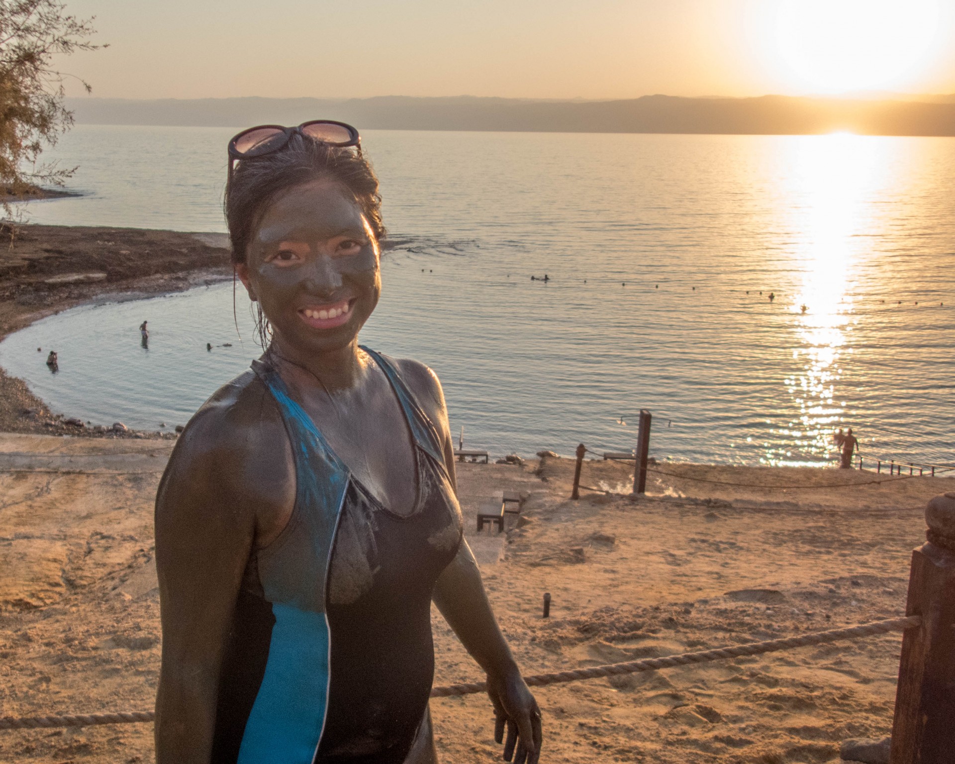 Woman covered in Dead Sea mud stands in front of the Sea at sunset at the Kempisnki Hotel