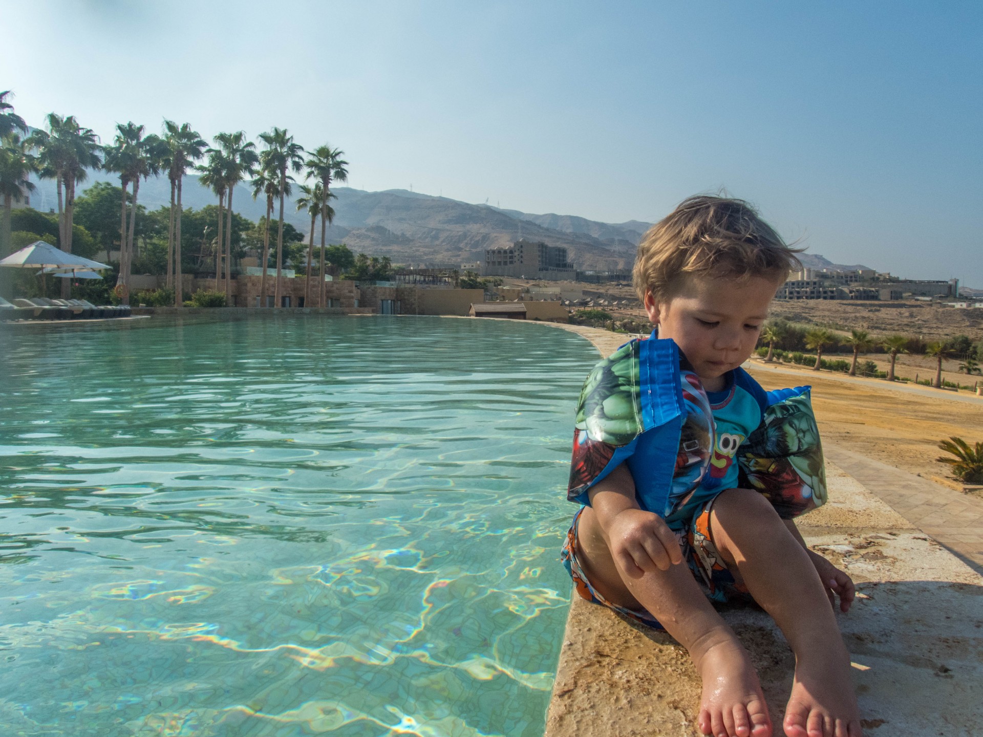 Toddler with water wings sits on the ledge of a pool with desert and palm trees in the background at the Kempinski Hotel
