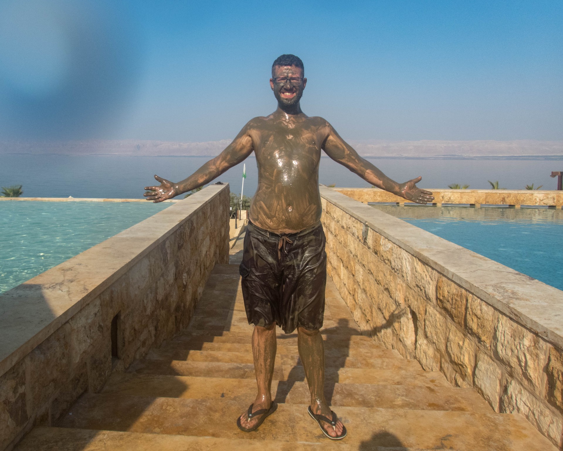Smiling man covered in mud holds his arms out in front of the Dead Sea at the Kempisnki Hotel