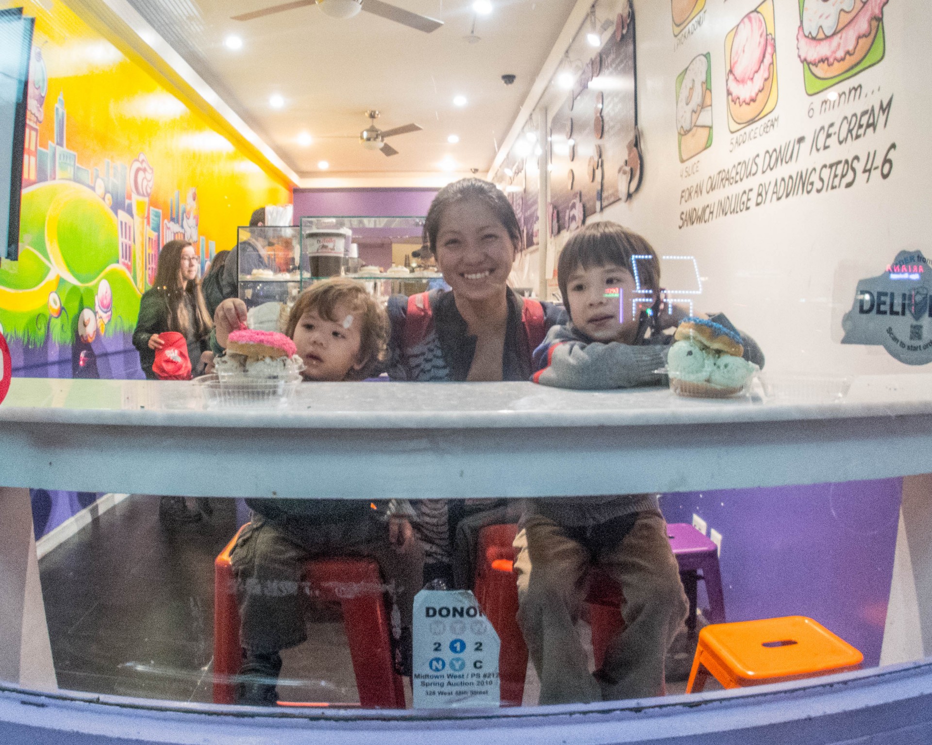 A mother and two young boys smile through the window of a bakery