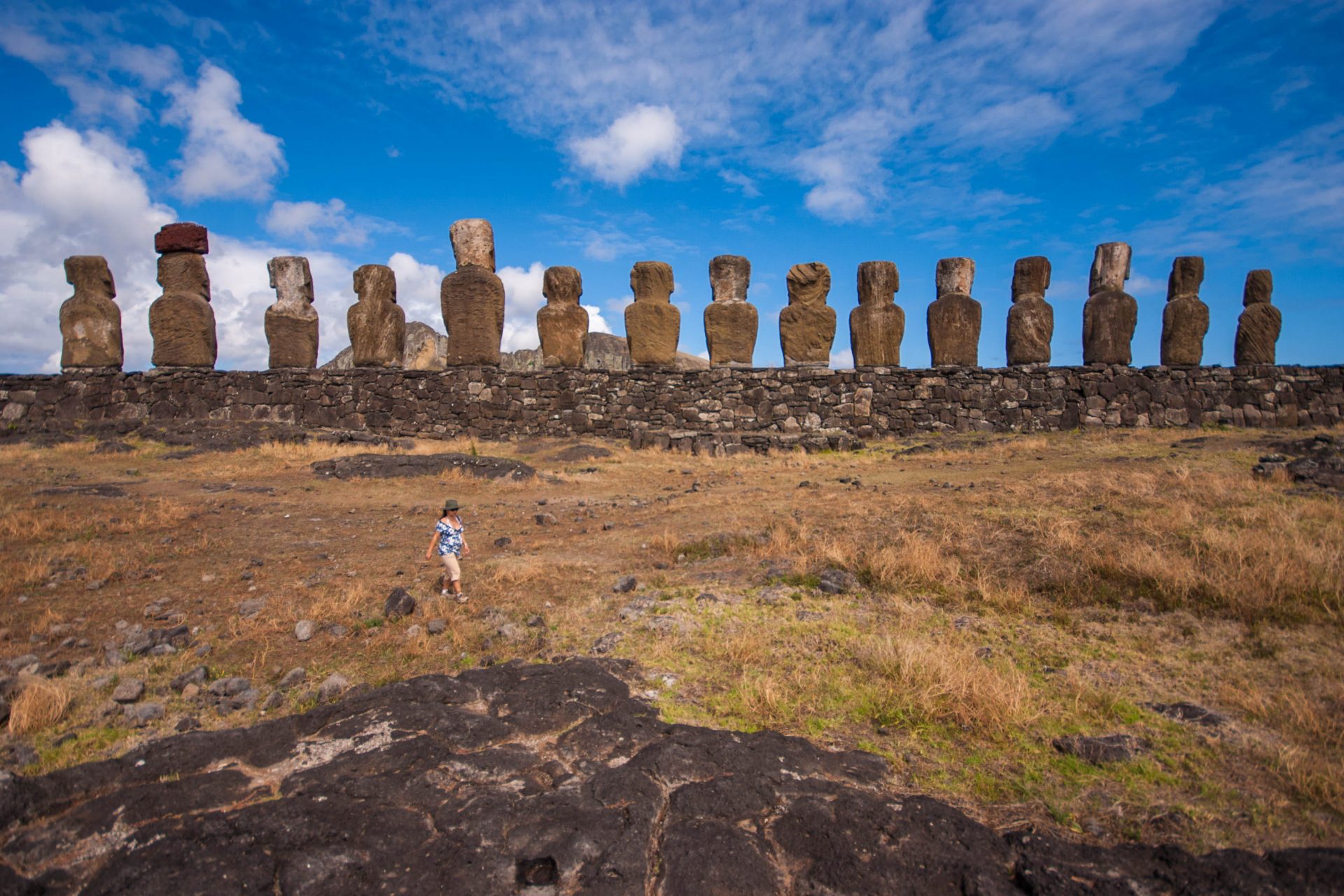 15 moai sit on an ahu looking inland towards Rano Raraku. In the foreground a pregnant woman walks towards the camera, dwarfed by the sculptures