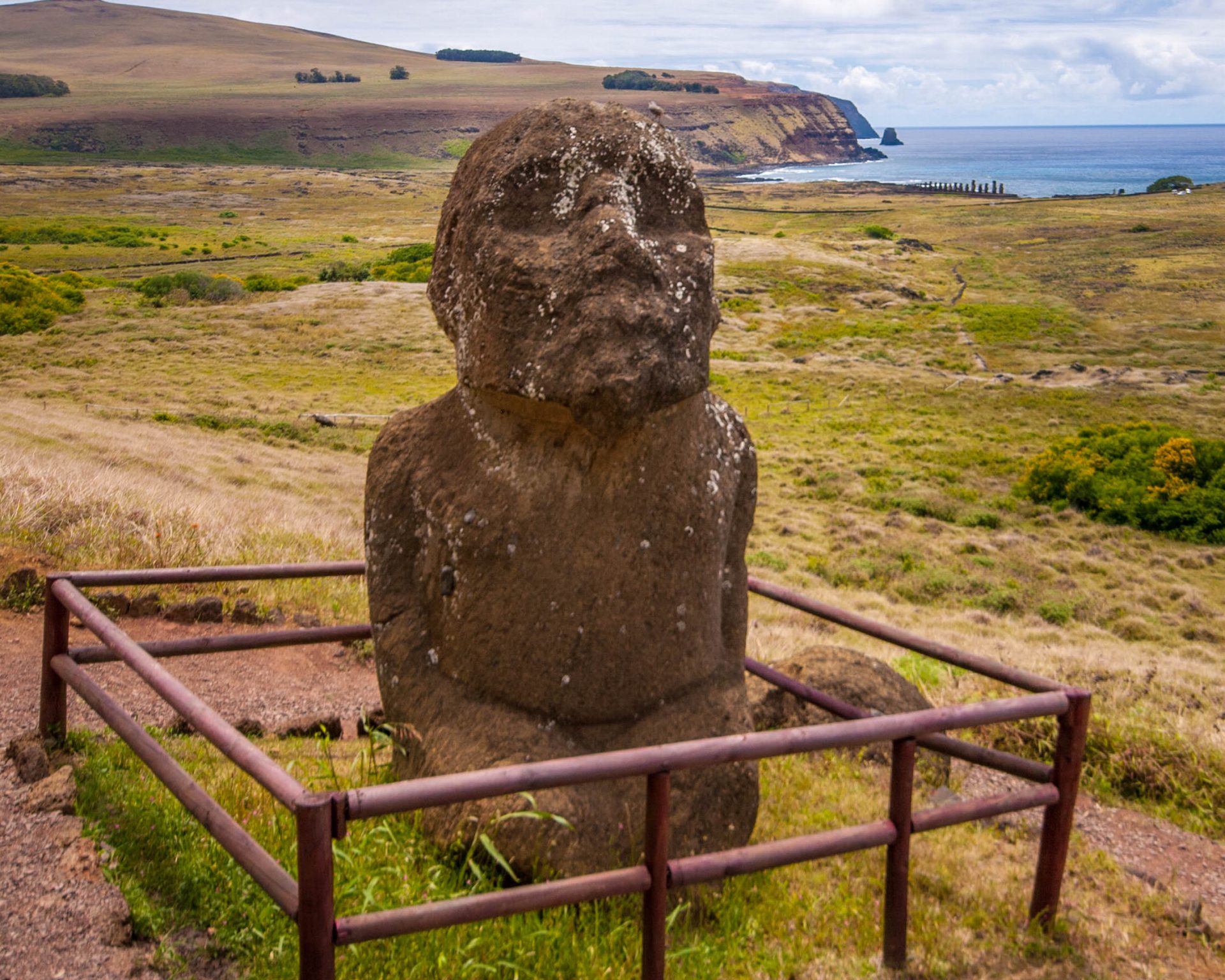 A unique moai sits in a fenced off area on a mountainside. The moai is kneeling and has a beard. In the background an ahu with 15 moai can be seen.