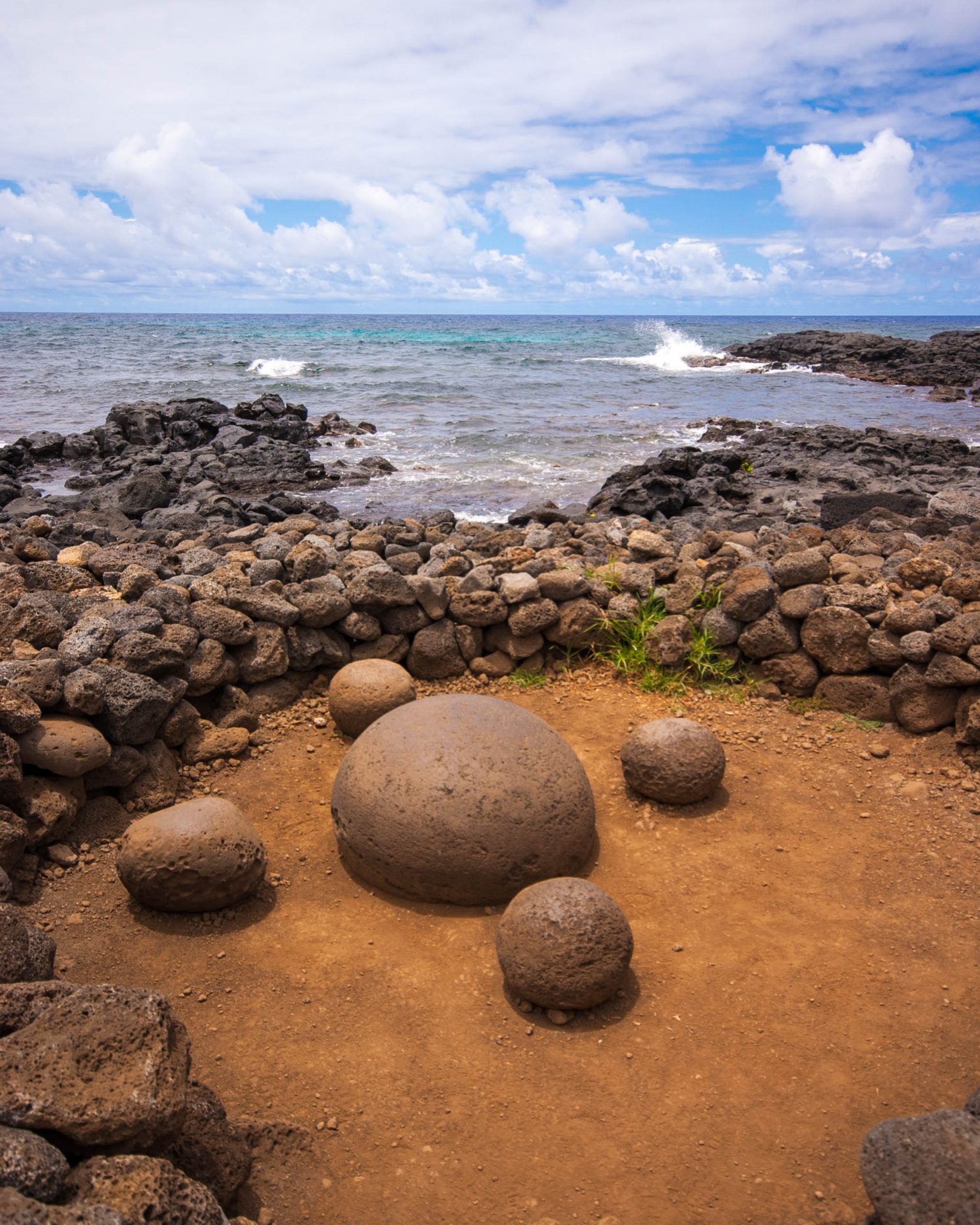 A large round rock is circled by four smaller round rocks sitting on red ground within a stone foundation. Blue skies and a wavy ocean are in the background
