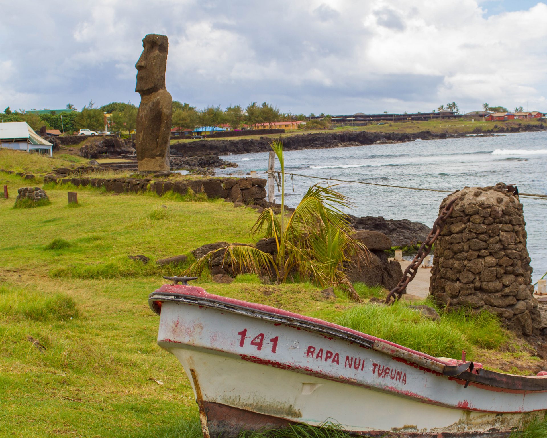 A small fishing boat sits on the green grass next to the ocean with a moai sculputre and a small town in the background