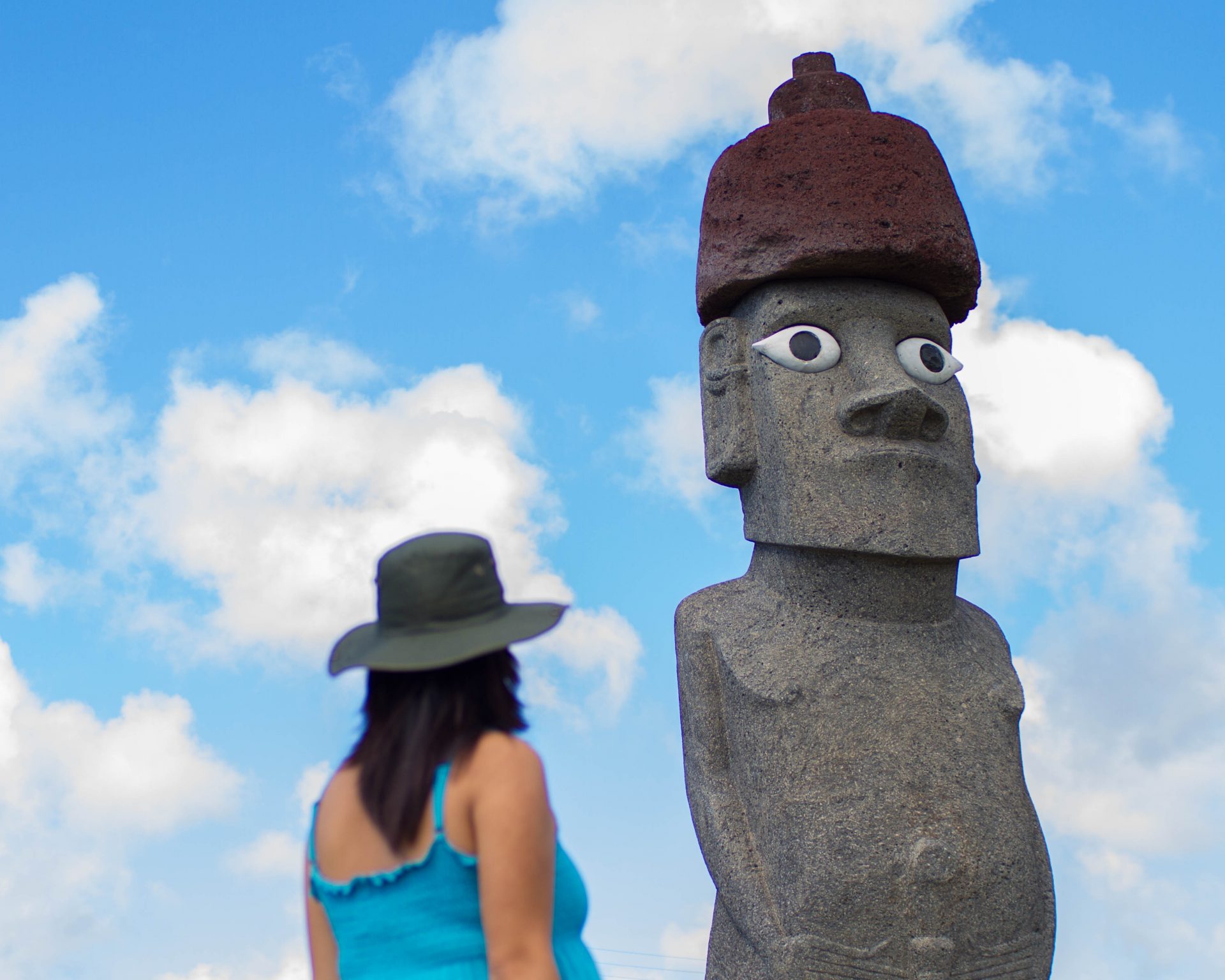 Woman wearing a green hat and blue shirt looks up at a completed replica sculpture of a moai backed with a blue sky on Easter Island