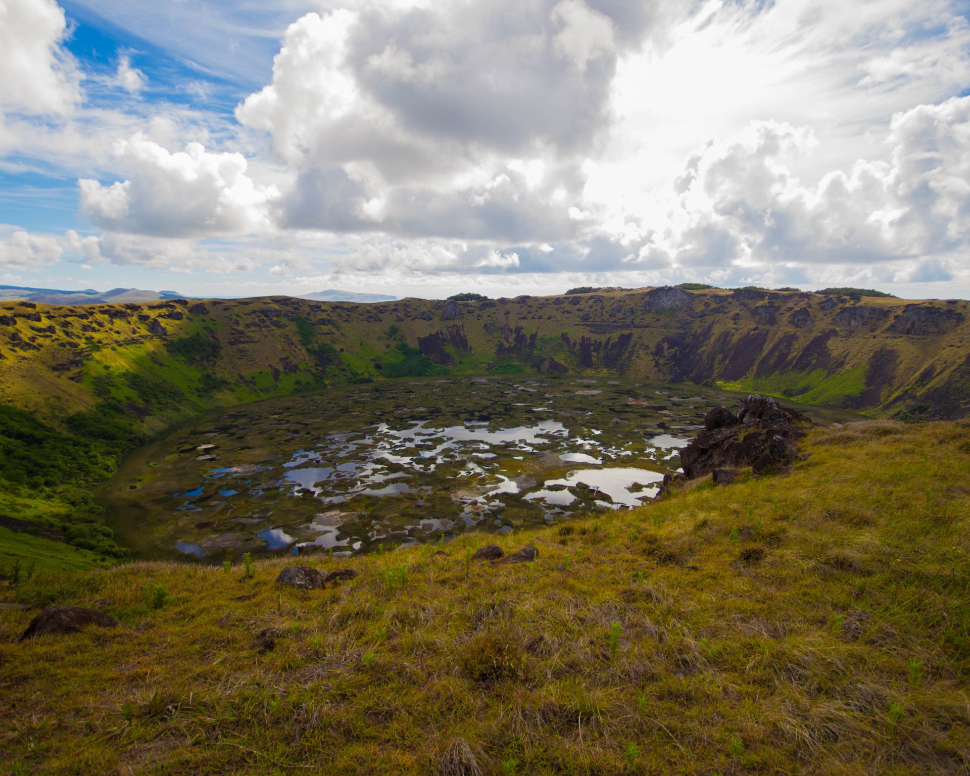 A colourful marsh reflects the clouds within a volcanic crater surrounded by lush green cliffs