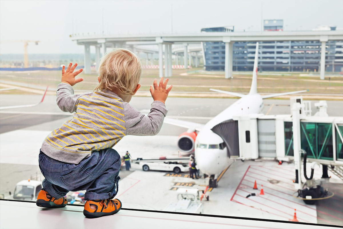 Toddler looking out the window of an airport as a plane is loaded in the background - Flying with a baby