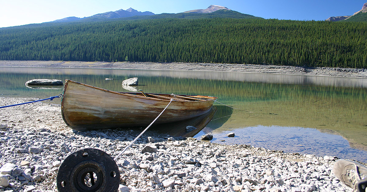 A wooden canoe anchored by a metal pipe sits in a crystal clear lake with forest and mountains in the background - Bucket list destinations in Canada