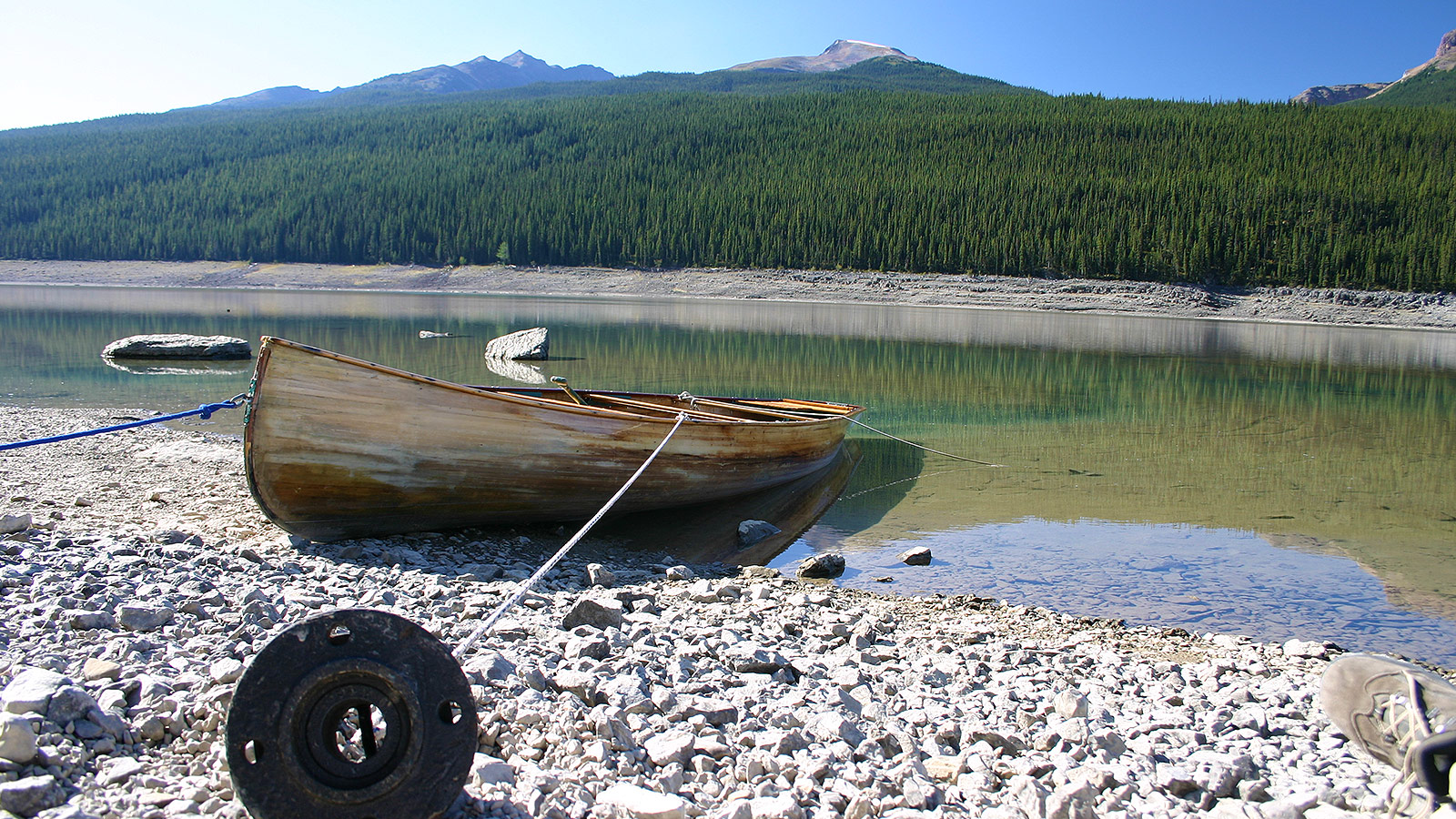 A wooden canoe anchored by a metal pipe sits in a crystal clear lake with forest and mountains in the background - Bucket list destinations in Canada