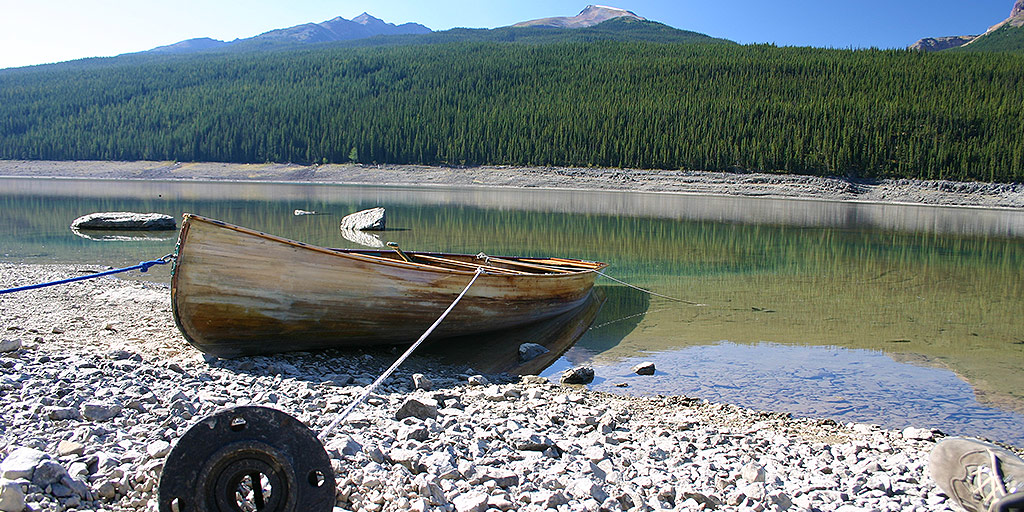 A wooden canoe anchored by a metal pipe sits in a crystal clear lake with forest and mountains in the background - Bucket list destinations in Canada