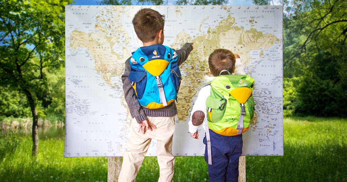 Two young boys in sweaters wearing cute bird backpacks stand in a forest and point at places on a world map