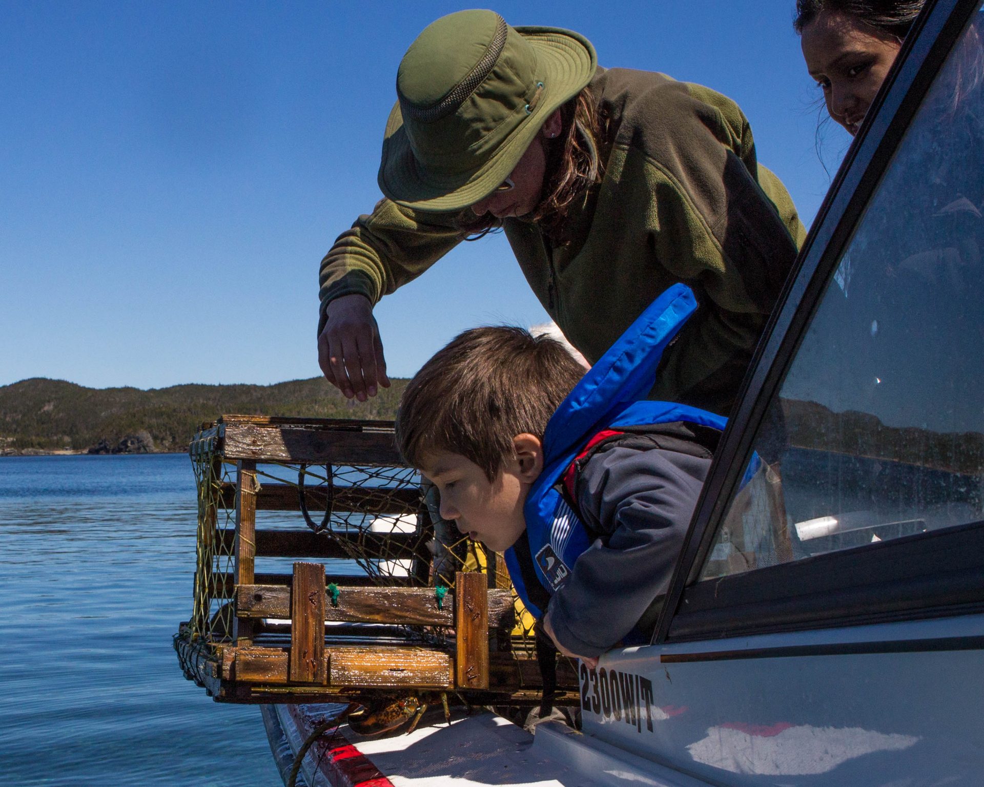 A woman wearing a hat checks a lobster trap on a boat while a young boy wearing a life jacket looks on - Icebergs in Twillingate