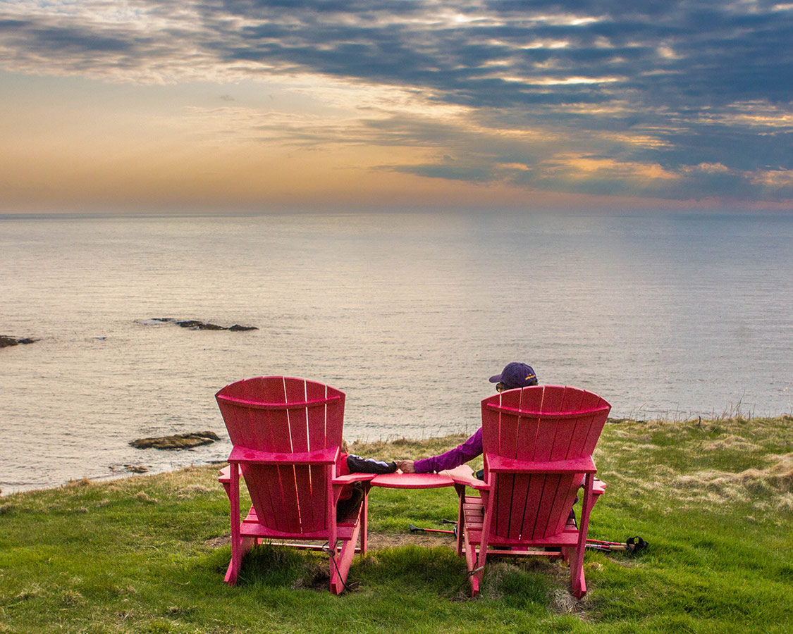 A woman and a toddler hold hands in large red muskoka chairs while watching the sunset at the top of a cliff overlooking the ocean - Hiking Green Gardens in Gros Morne National Park