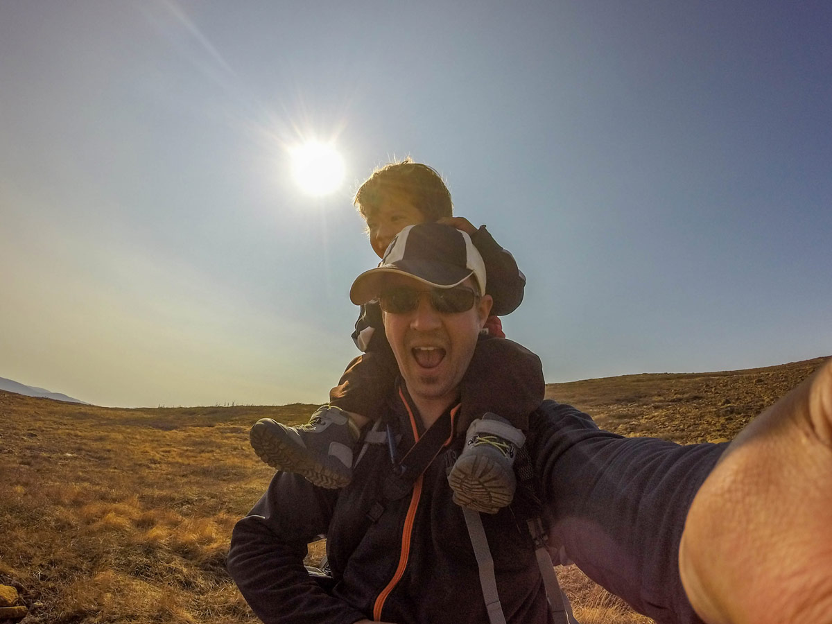 A father smile at the camera while giving a piggy back to a toddler on a hike through Gros Morne National Park - Hiking Green Gardens in Gros Morne National Park
