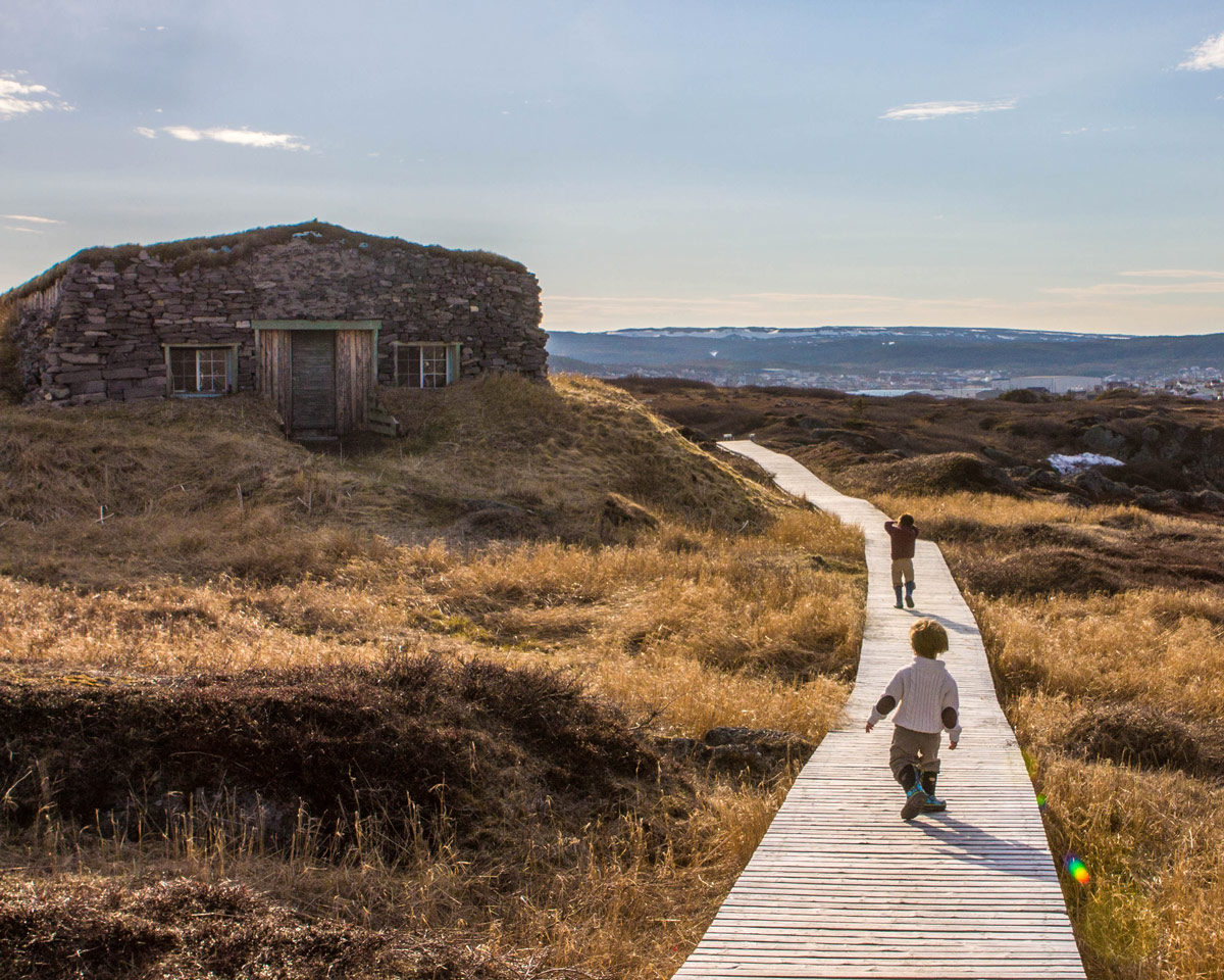 Two young boys walk along a boardwalk towards a grass covered stone building - Newfoundland Viking Trail