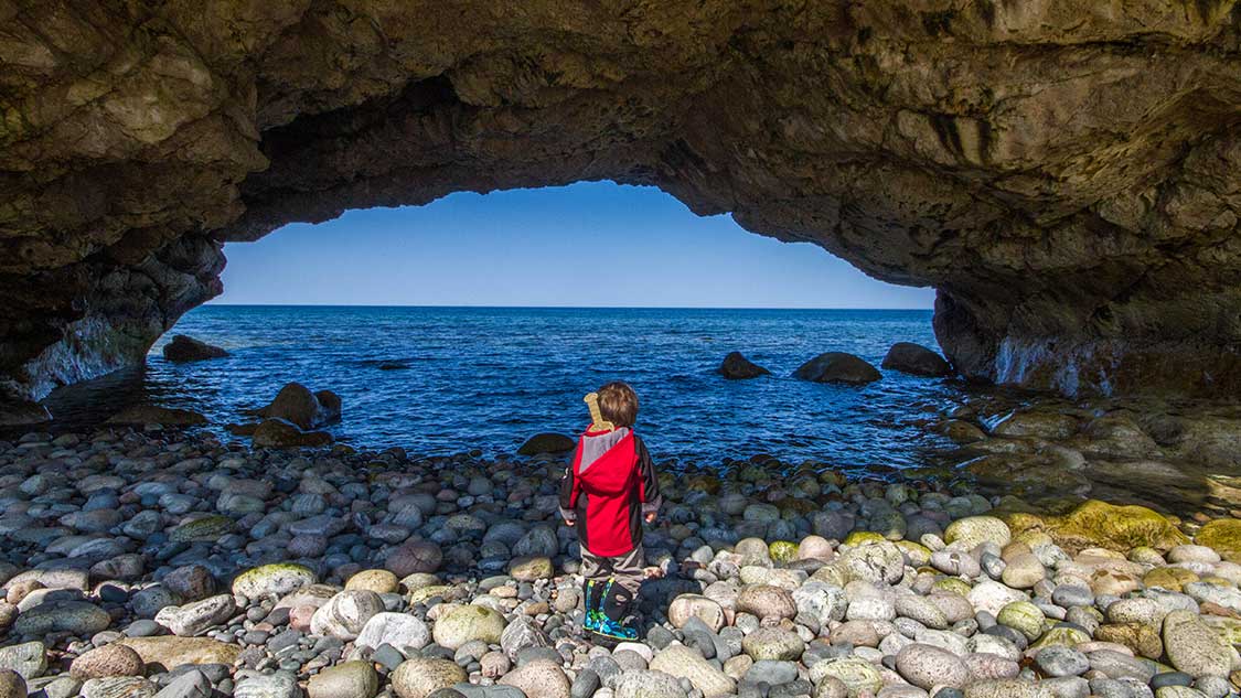 Boy with a viking sword stands on a rocky beach looking through a sea cave on the Newfoundland Viking Trail