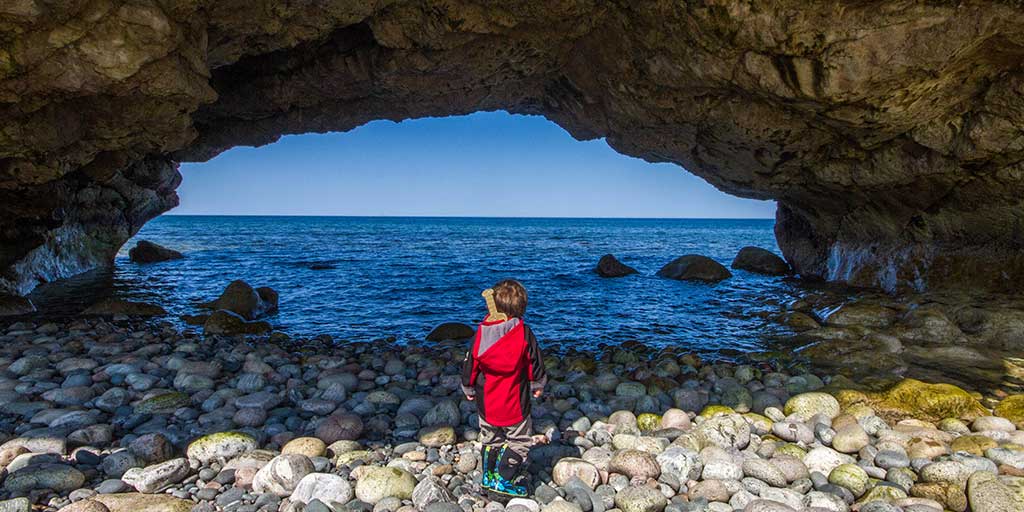 Boy with a viking sword stands on a rocky beach looking through a sea cave on the Newfoundland Viking Trail