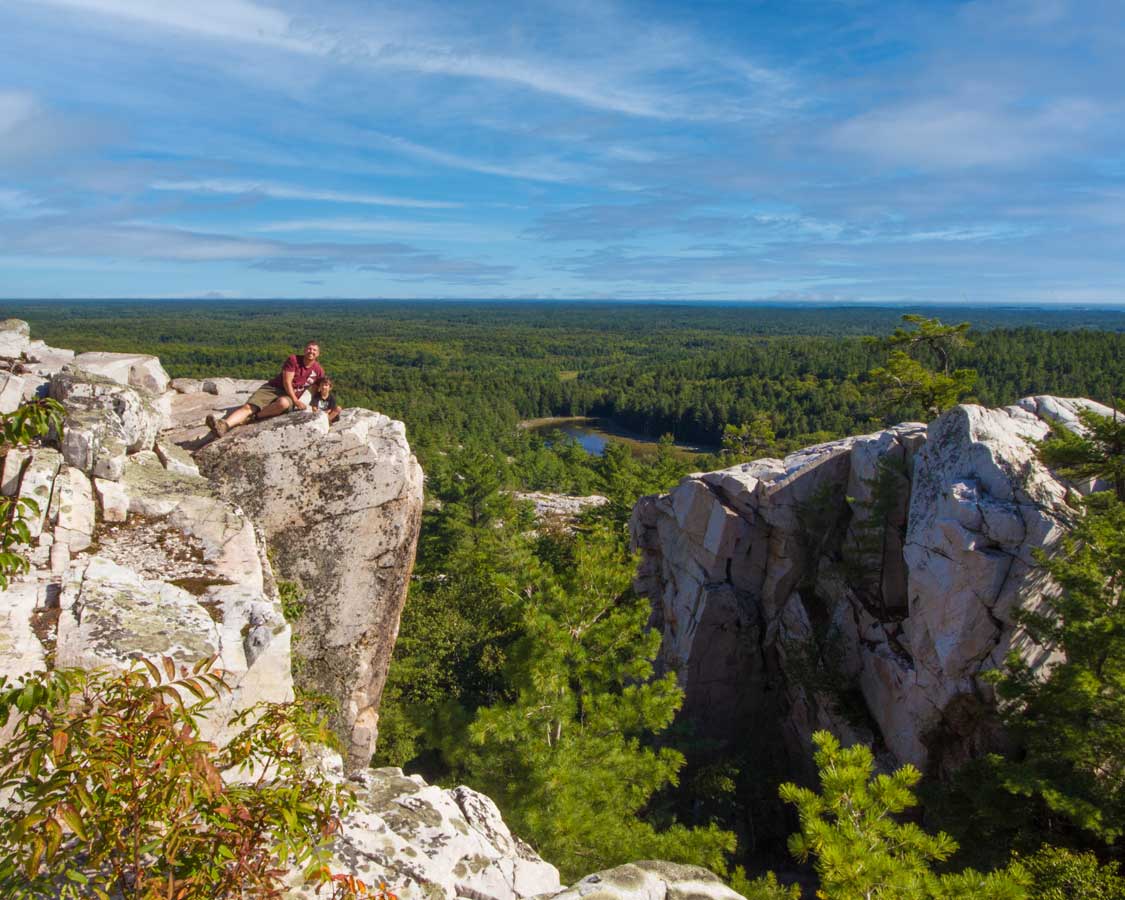 Father and son pose for a photo at the top of a rocky cliff overlooking forests in Killarney Provincial Park