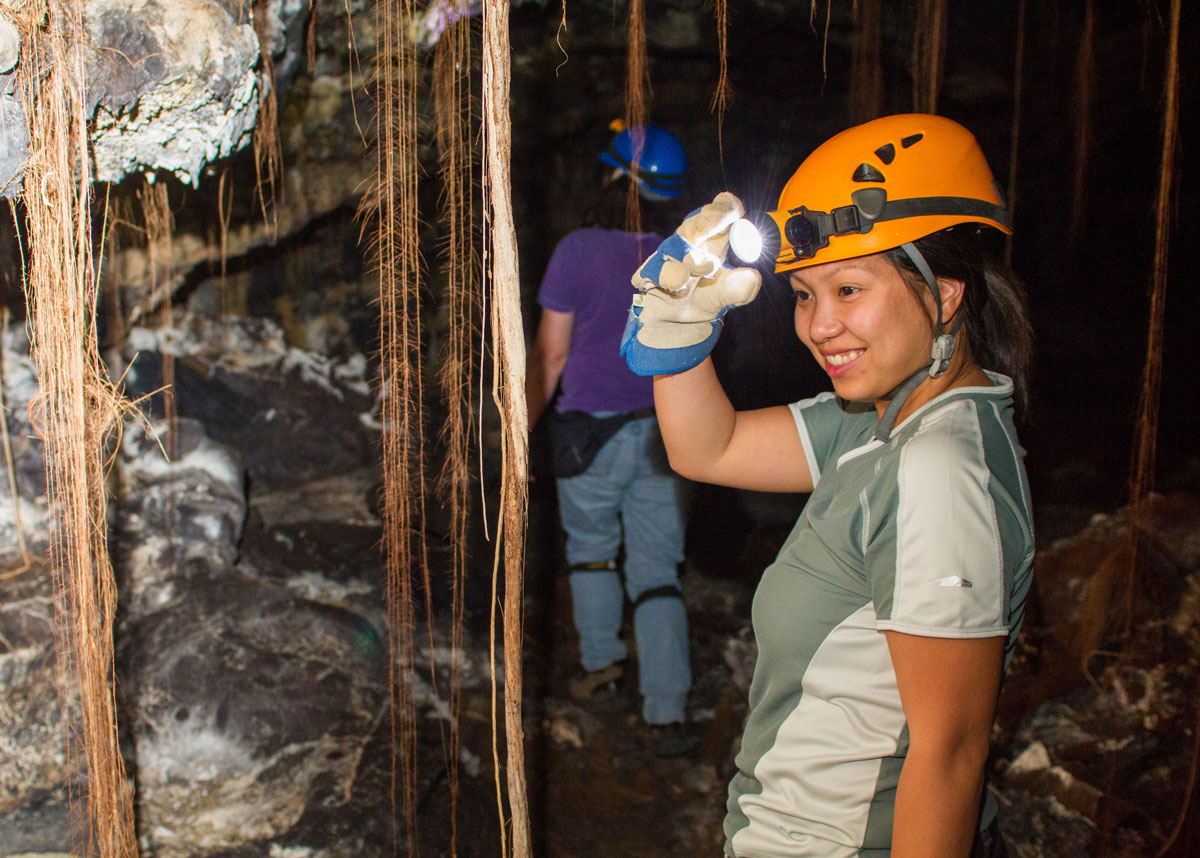 A woman wearing a hardhat and headlamp looks at vegetation in a lava tube - caves you can visit with kids