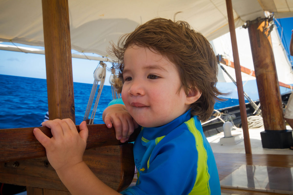 A young boy with wind blowing though his brown hair looks over the bow of a sail boat at sea - Swimming with turtles in the Tobago Cays