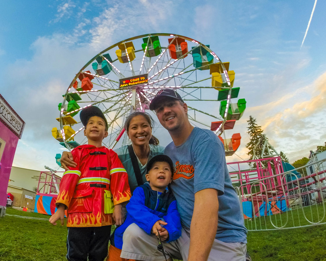 A young family smiling at the camera in front of a colourful ferris wheel while attending a fall fair, one of the 5 kid-friendly fall activities in Ontario.