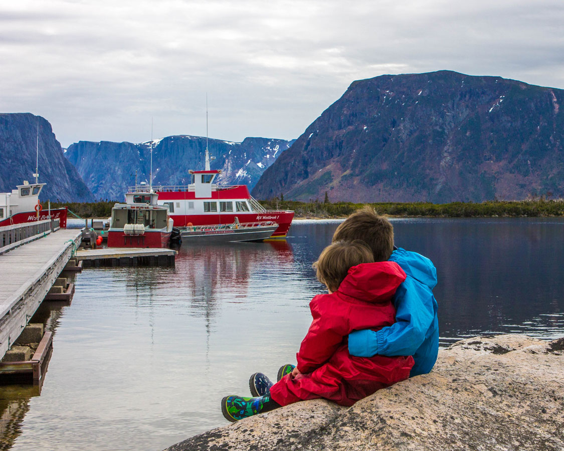Two young boys wearing rain outfits hug in front of a marina at Western Brook Pond in Gros Morne National Park