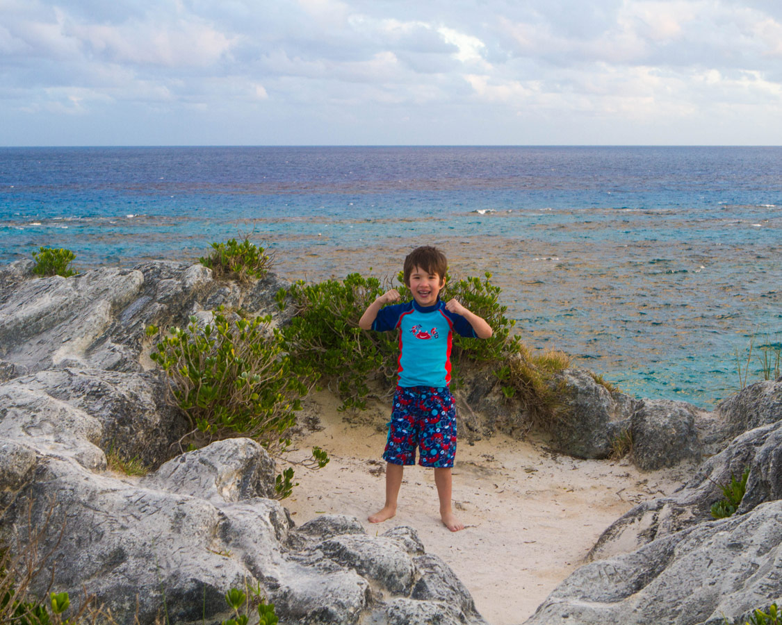 A young boy flexes his muscles on a patch of sand at the top of a cliff overlooking the ocean in Bermuda