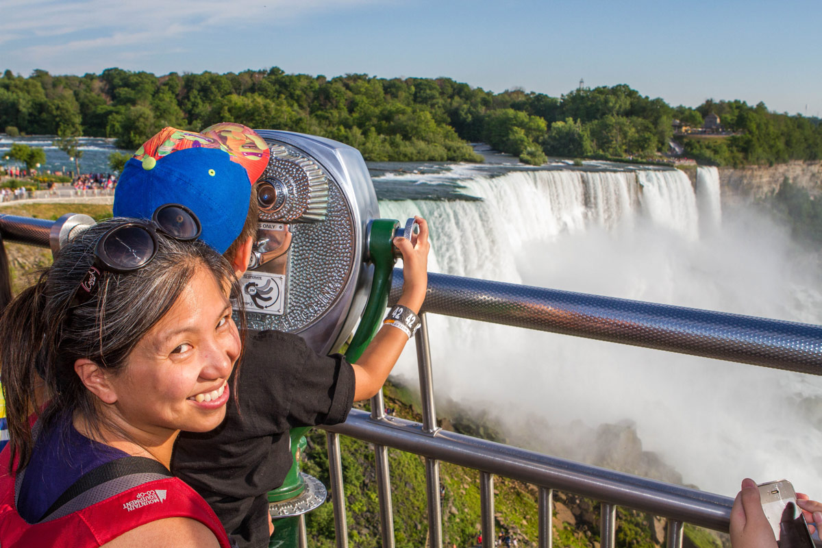 A woman smiles at the camera while her son looks through viewpoint binoculars at Niagara Falls New York