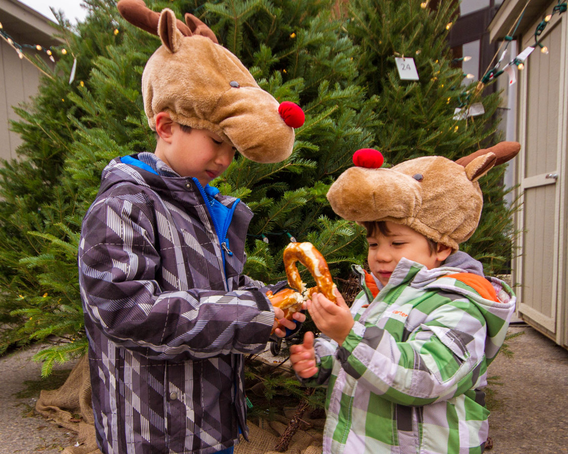 Two young boys wearing reindeer hats eat a pretzel in front of a christmas tree at the Christkindle Christmas Market in Kitchener, Ontario