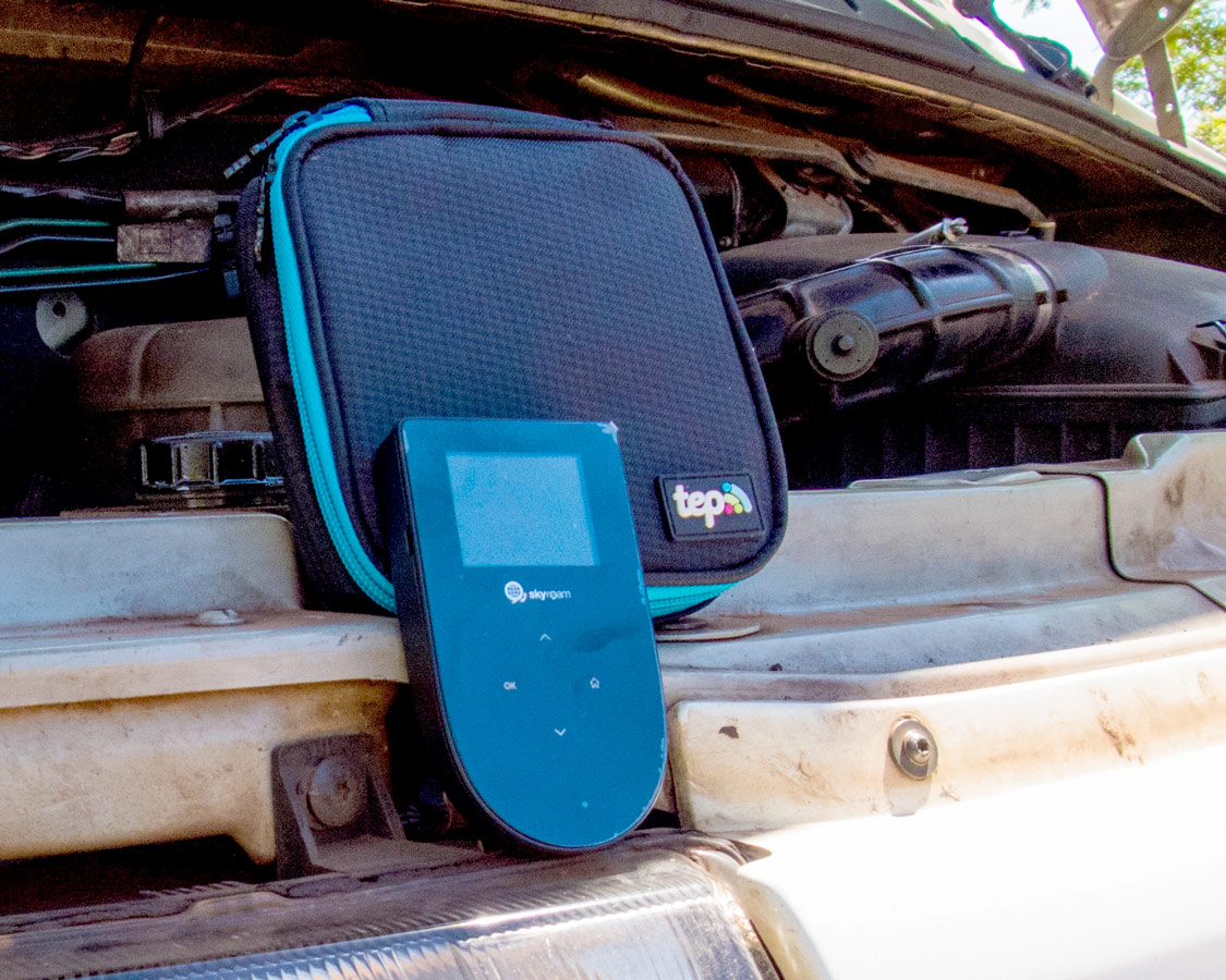Small black digital Tep wifi hotspot device and carrying case sitting on the open hood of a van that has broken down.