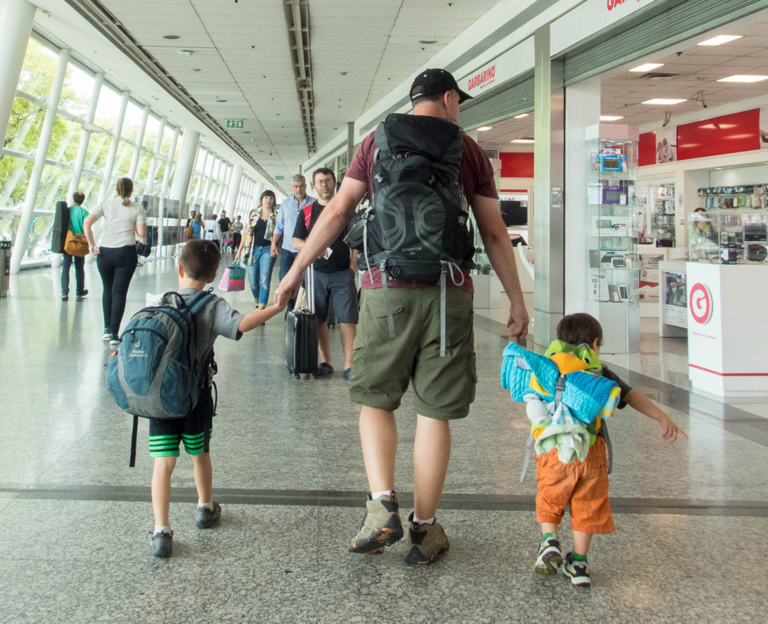 A father and his two young sons walk through an airport while carrying their backpackson a family travel journey